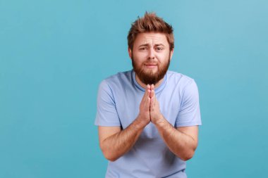 Please, I'm begging. Portrait of bearded man holding arms in prayer, asking help or forgiveness with imploring eyes, sincere asking permission. Indoor studio shot isolated on blue background.