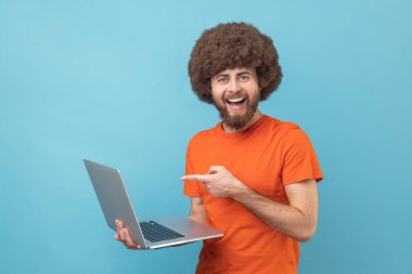 Portrait of man with Afro hairstyle wearing orange T-shirt pointing laptop screen and looking at camera with happy expression, toothy smile. Indoor studio shot isolated on blue background.