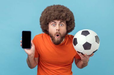 Portrait of shocked man with Afro hairstyle wearing T-shirt standing with soccer ball and showing cell phone with empty display for advertisement. Indoor studio shot isolated on blue background.