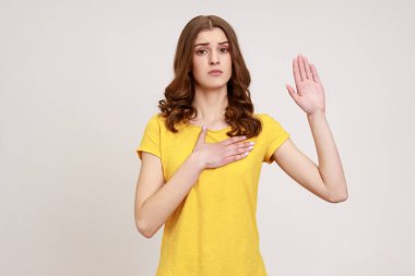 Serious conscious brown haired woman of young age holding one hand on chest and raising another swearing, trust and honest, patriotism. Indoor studio shot isolated on gray background.