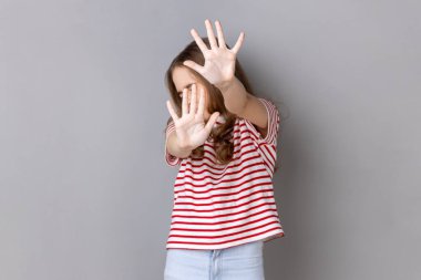 Portrait of scared little dark haired girl wearing striped T-shirt gesturing stop with palms and looking turning away, being frighten. Indoor studio shot isolated on gray background.