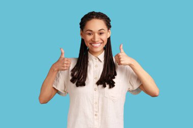Great deal. Good looking woman with black dreadlocks smiling happily, keeps thumbs up, approves something, has cheerful expression, wearing white shirt. Indoor studio shot isolated on blue background.