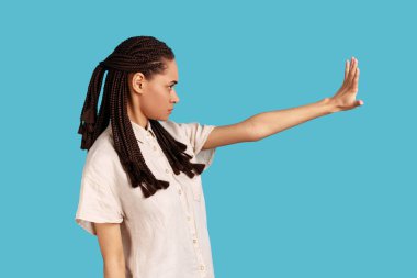 Side view portrait of serious woman with black dreadlocks keeps palm in stop gesture, asks not to bother her, looks angrily ahead, wearing white shirt. Indoor studio shot isolated on blue background.