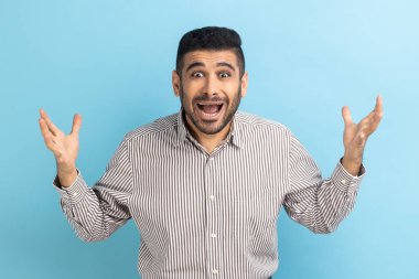 Irritated annoyed businessman with beard has problems, gesticulating with hands, crisis and negativity, misunderstanding, wearing striped shirt. Indoor studio shot isolated on blue background.