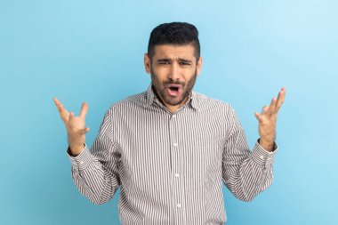 Annoyed stressed businessman with beard asking what do you want and spreading hands, family quarrel, misunderstanding, wearing striped shirt. Indoor studio shot isolated on blue background.