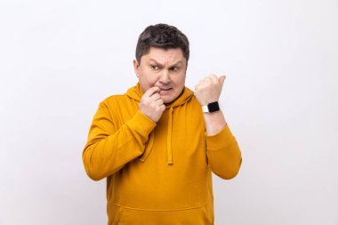 Nervous middle aged man biting finger nails and looking at his wrist clock with anxious, worried about deadline, wearing urban style hoodie. Indoor studio shot isolated on white background.