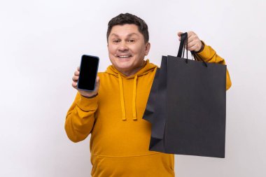 Satisfied man holding cell phone and shopping bags, advertising online store on mobile device, good delivery application, wearing urban style hoodie. Indoor studio shot isolated on white background.