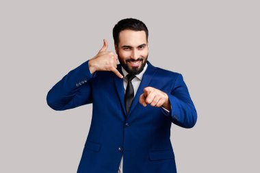 Delighted bearded man standing with telephone hand gesture and smiling to camera, flirting offering to contact by phone, pointing finger to camera. Indoor studio shot isolated on gray background.