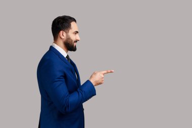 Side view of bearded man pointing finger away with toothy smile on face, empty space for your advertisement, wearing official style suit. Indoor studio shot isolated on gray background.