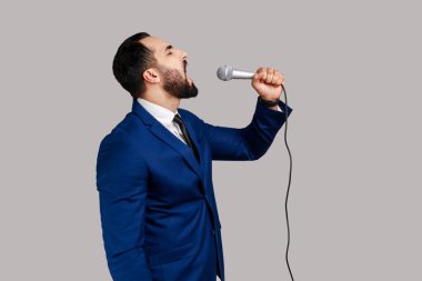 Side view of bearded man singing songs, holding microphone, singer making performance, keeps eyes closed, wearing official style suit. Indoor studio shot isolated on gray background.