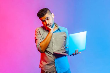 Portrait of bored sleepy man in shirt looking at camera with indifferent expression, having tedious online conversation. Indoor studio shot isolated on colorful neon light background.