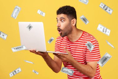 Money falling and shocked rich man. Money rain and portrait of shocked man holding laptop and looking at laptop display with big shocked unbelievable eyes. Indoor isolated on yellow background