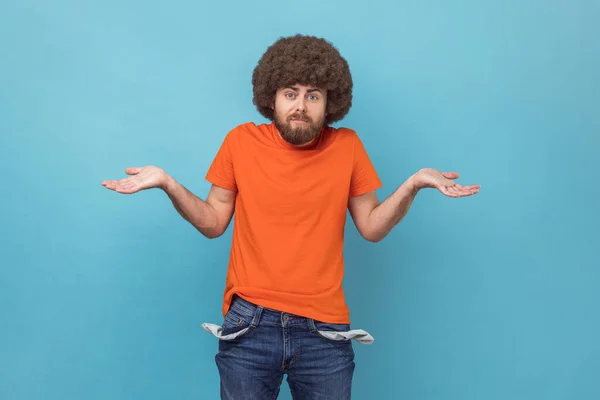 Portrait of man with Afro hairstyle wearing orange T-shirt showing empty pockets and looking frustrated about loans and debts, has no money, jobless. Indoor studio shot isolated on blue background.