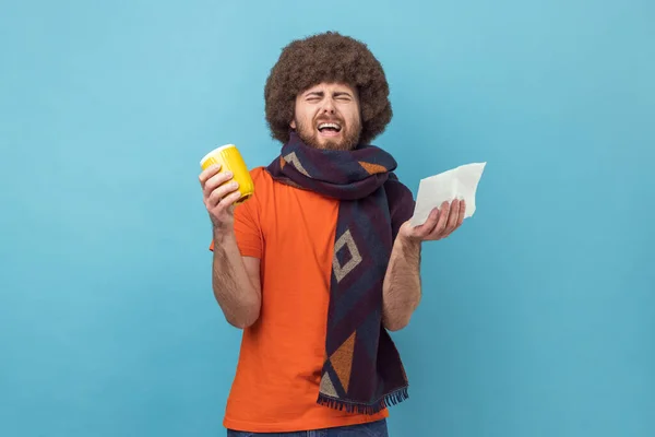 Portrait of man with Afro hairstyle in orange T-shirt holding mug and tissue for runny nose, taking medicine with hot tea, suffering fever, headache. Indoor studio shot isolated on blue background.