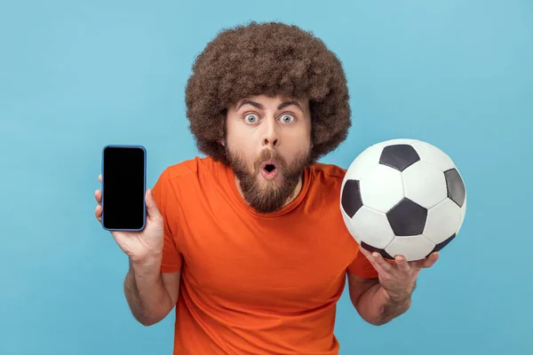 Portrait of shocked man with Afro hairstyle wearing T-shirt standing with soccer ball and showing cell phone with empty display for advertisement. Indoor studio shot isolated on blue background.
