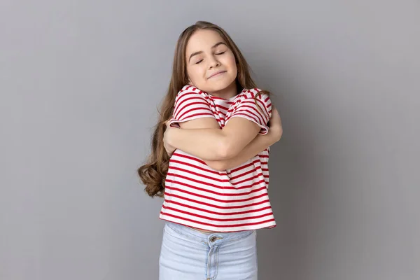 I love myself. Portrait of self-satisfied egoistic little girl wearing striped T-shirt embracing herself and smiling with pleasure, feeling self-pride. Indoor studio shot isolated on gray background.