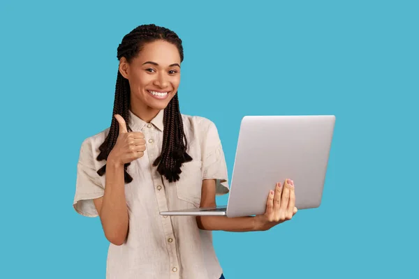 Smiling cheerful woman with dreadlocks using laptop, raising her thumb up, happy and satisfied with great news on his online trade, wearing white shirt. Indoor studio shot isolated on blue background.