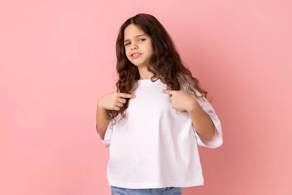 Portrait of self-confident narcissistic little girl wearing white T-shirt standing pointing himself, feeling self-important, proud, famous. Indoor studio shot isolated on pink background.