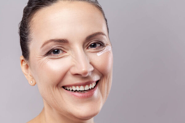 Closeup portrait of beautiful middle aged woman with perfect face skin and cosmetic cream near eyes, looking at camera with toothy smile. Indoor studio shot isolated on gray background.