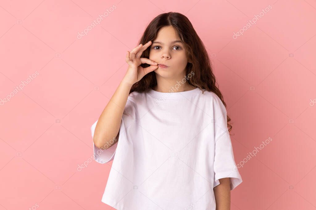 Portrait of charming little girl wearing white T-shirt with mystery look making zip gesture to close mouth, keeping secret, zipping lips. Indoor studio shot isolated on pink background.
