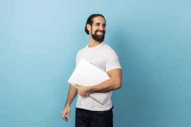 Side view of positive man with beard wearing white T-shirt standing, holding closed laptop or folder, looking back with optimistic expression. Indoor studio shot isolated on blue background.