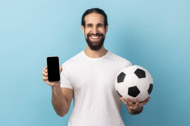 Portrait of happy man with beard wearing white T-shirt holding soccer ball and smartphone empty black display, ticket booking for championship. Indoor studio shot isolated on blue background.