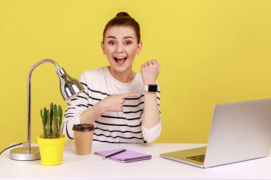 Look at clock, time to work. Positive hard working woman sitting workplace and pointing wrist watch, looking at camera, rejoice to start work. Indoor studio studio shot isolated on yellow background.