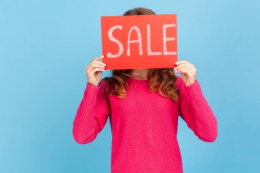 Portrait of unknown woman wearing pink pullover, covering face with Sale inscription, market discounts, black friday, low prices. Indoor her studio shot isolated on blue background.
