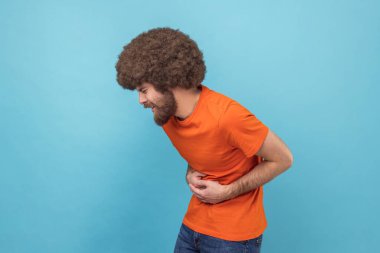 Stomach pain. Side view of sick sad unhealthy man with Afro hairstyle wearing orange T-shirt standing and holding his painful belly. Indoor studio shot isolated on blue background.