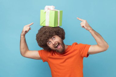 Portrait of positive funny man with Afro hairstyle wearing orange T-shirt holding gift box on his head and pointing with fingers, expressing excitement. Indoor studio shot isolated on blue background.
