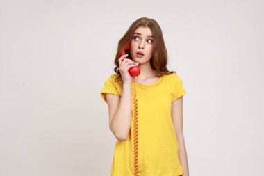 Portrait of amazed young curly haired female in yellow casual style T-shirt talking vintage phone, surprised by conversation, looking away. Indoor studio shot isolated on gray background.