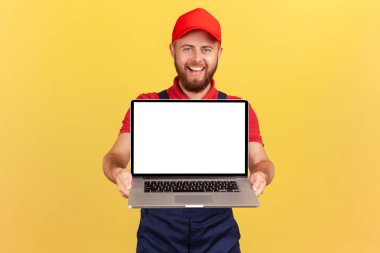 Portrait of delighted handsome worker man in red cap holding laptop, showing blank screen with copy space for advertisement and promotional text. Indoor studio shot isolated on yellow background.