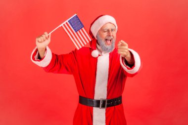 Satisfied elderly man with gray beard wearing santa claus costume waving USA flag winking and pointing to camera, keeps mouth open. Indoor studio shot isolated on red background.