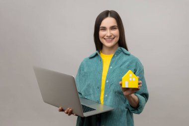 Woman real estate agent standing with paper house and laptop in hand, helping with rent, working on project online, wearing casual style jacket. Indoor studio shot isolated on gray background.
