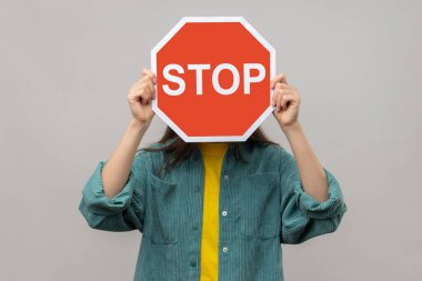 Attention, prohibited way. Unknown person hiding face behind Stop symbol, red traffic sign warning of restricted access, banned service, wearing casual style jacket. Indoor studio shot isolated on gray background.