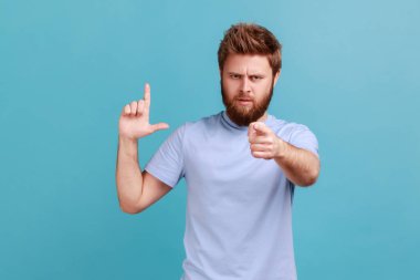 Portrait of disrespectful rude bearded man showing loser gesture and pointing on you, abuser, blaming for mistake and dismissing from job. Indoor studio shot isolated on blue background.