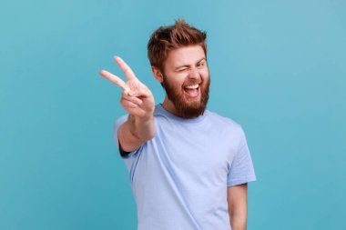 Portrait of happy optimistic bearded man doing victory gesture and winking playfully to camera, showing peace, v sign with double fingers. Indoor studio shot isolated on blue background.