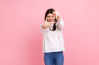 Smiling woman looking through finger frame, imitating to take photo, focusing interesting moment, wearing white casual style sweater. Indoor studio shot isolated on pink background.