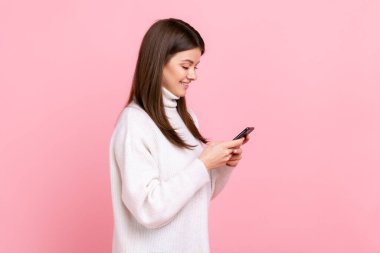 Side view of portrait optimistic girl using smartphone with happy expression, hearing pleasant news, wearing white casual style sweater. Indoor studio shot isolated on pink background.