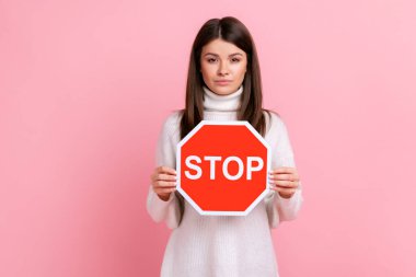 Serious brunette woman standing with calm strict facial expression, shows red stop symbol to camera, wearing white casual style sweater. Indoor studio shot isolated on pink background.