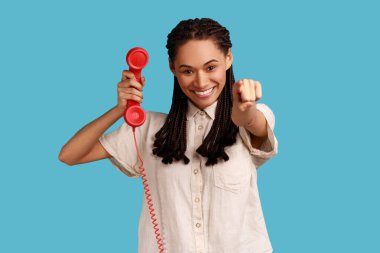 Beautiful woman with black dreadlocks pointing finger to camera, holding handset of red vintage landline phone, answering calls, wearing white shirt. Indoor studio shot isolated on blue background.