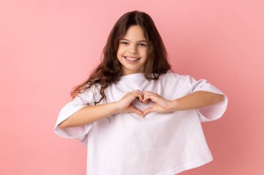 Portrait of little girl wearing white T-shirt standing with raised hands and making heart or love gesture and looking at camera with toothy smile. Indoor studio shot isolated on pink background.