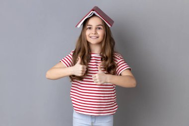 Portrait of delighted little girl in striped T-shirt covering head with book and smiling to camera, child having fun, showing thumbs up. Indoor studio shot isolated on gray background.