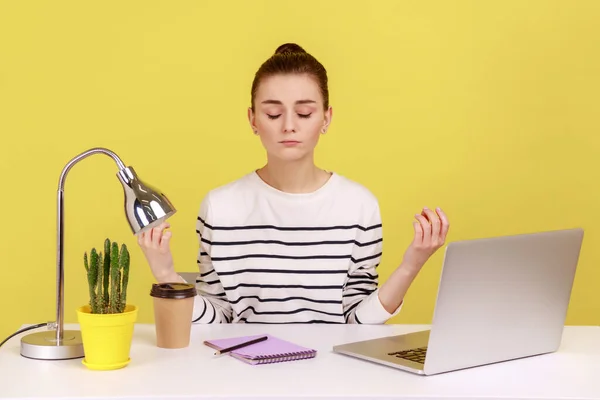 Peaceful mind, break at work. Calm woman sitting at workplace with laptop and raising hands in mudra gesture, meditating resting at home office. Indoor studio studio shot isolated on yellow background