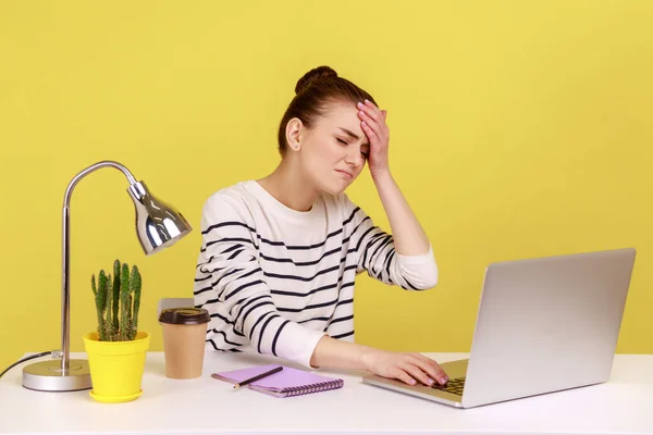 Forgetful woman manager sitting at workplace with laptop and keeping hand on forehead, blaming herself with facepalm gesture, expressing sorrow. Indoor studio studio shot isolated on yellow background