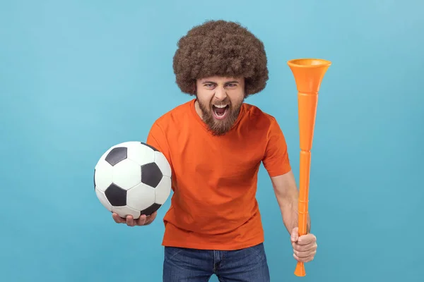 Portrait of man with Afro hairstyle wearing orange T-shirt standing with soccer ball and horn, screaming happily with crazy expression. Indoor studio shot isolated on blue background.