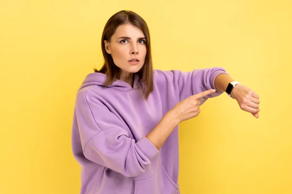 Portrait of bossy woman with dark hair pointing wrist watch and looking annoyed displeased, showing clock to hurry up, wearing purple hoodie. Indoor studio shot isolated on yellow background.