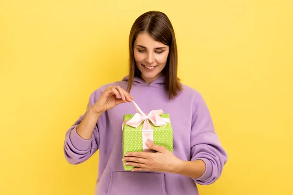Young beautiful smiling woman feeling happy, opens present box with happy satisfied expression, being glad to get gift, wearing purple hoodie. Indoor studio shot isolated on yellow background.