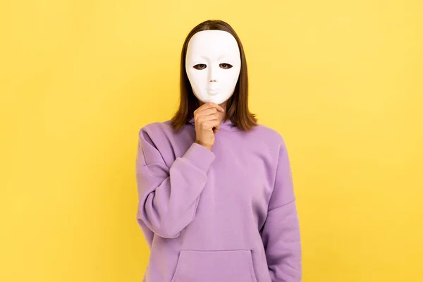 Portrait of anonymous dark haired woman covering her face with white mask, hiding personality, conspiracy and privacy, wearing purple hoodie. Indoor studio shot isolated on yellow background.