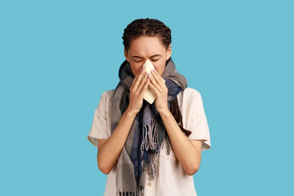 Portrait of ill woman with black dreadlocks sneezes in handkerchief, standing wrapped in scarf, catching cold, flu symptoms, wearing white shirt. Indoor studio shot isolated on blue background.